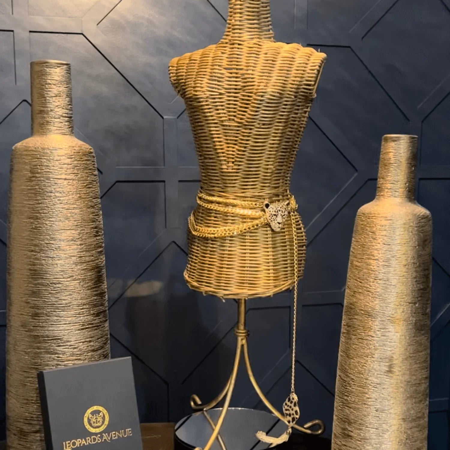 Wicker mannequin with gold belt, two tall gold vases, and Leopards Avenue box on dark textured wall
