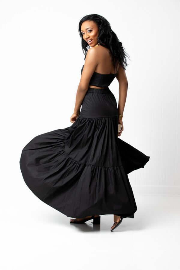 Woman posing in black strapless crop top and flowing maxi skirt against white background
